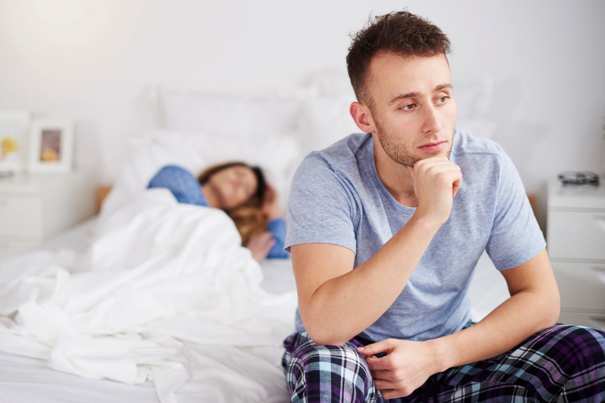 Man sitting at the end of the bed thinking while a woman sleeps behind him out of focus.