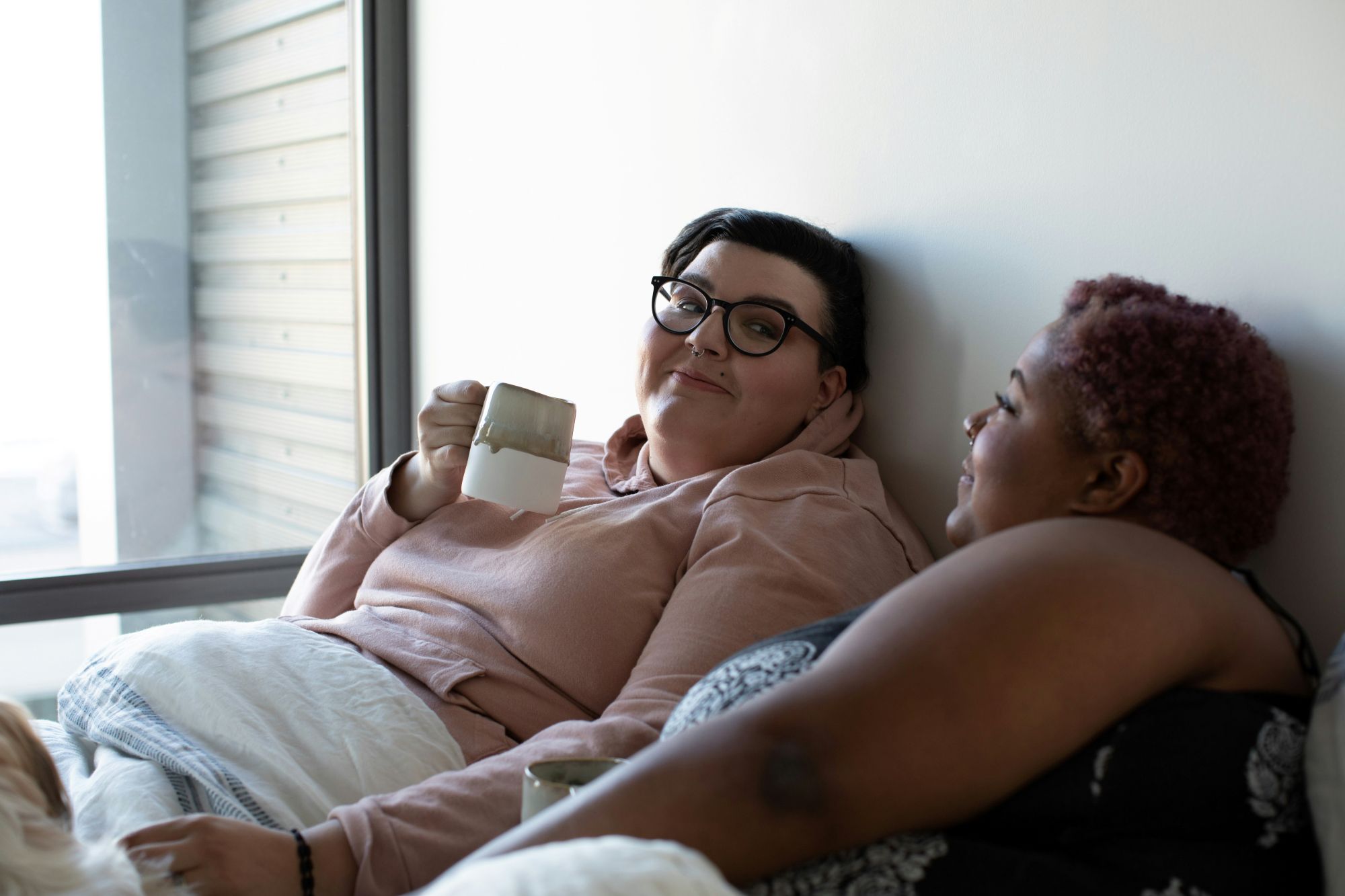 Two curvy women relaxing in bed together and looking at each other lovingly.