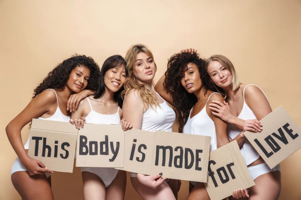 A group of 5 women standing together. They are different races and sizes, and they are each holding a sign with one of the words in the sentence This body is made for love