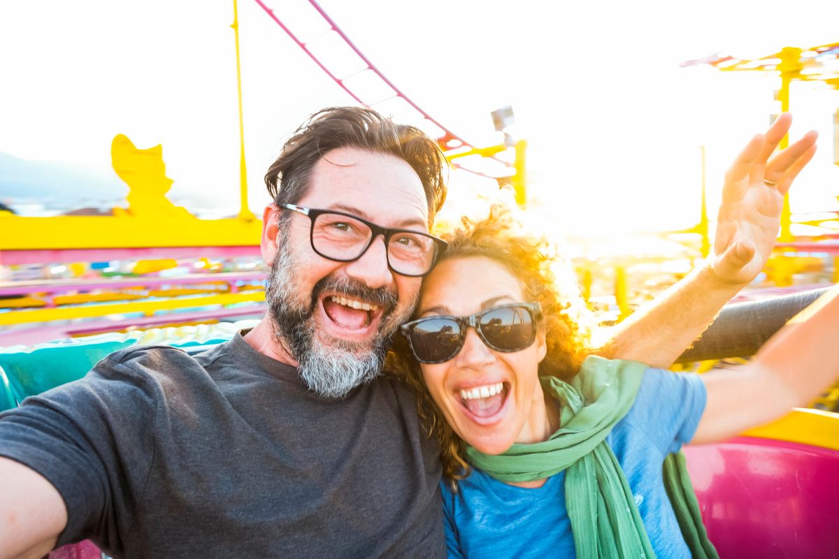 Man and woman smiling, seated with their arms up at the start of rollercoaster ride.